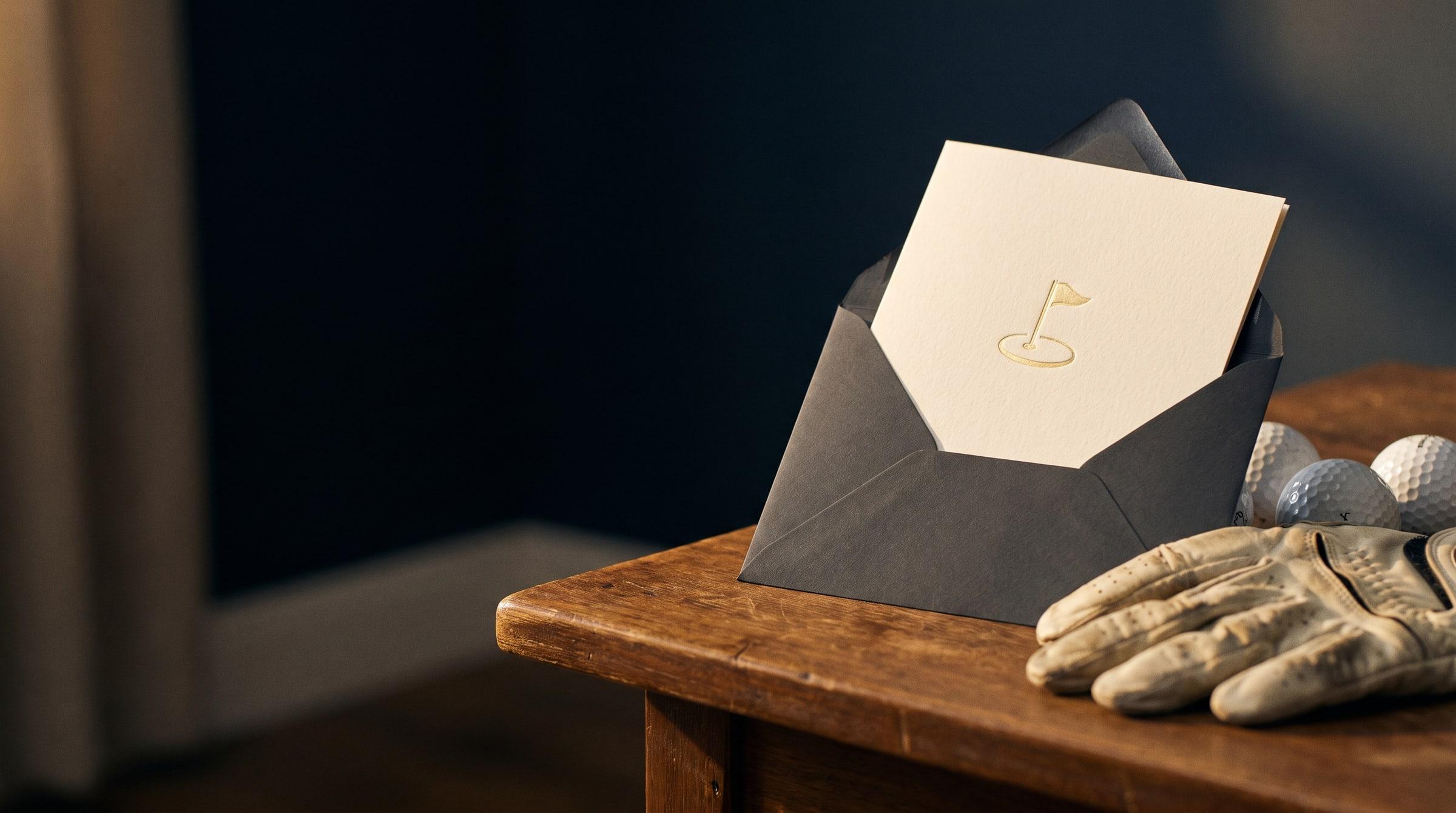 An ivory invitation with a gold golf-flag emblem rests beside a golf glove on a walnut table